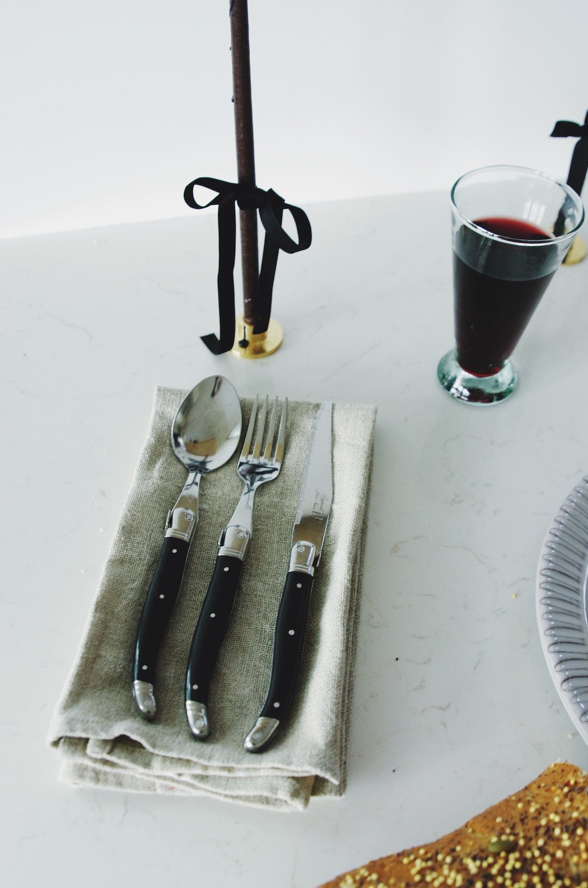 A set of elegant French Laguiole cutlery, including a spoon, fork, and two knives with black handles, is neatly placed on a natural linen napkin. To the right, there's a clear glass filled with red wine, and in the background, a candle with a black ribbon around its brass holder adds a touch of sophistication to the simple yet refined table setting.
