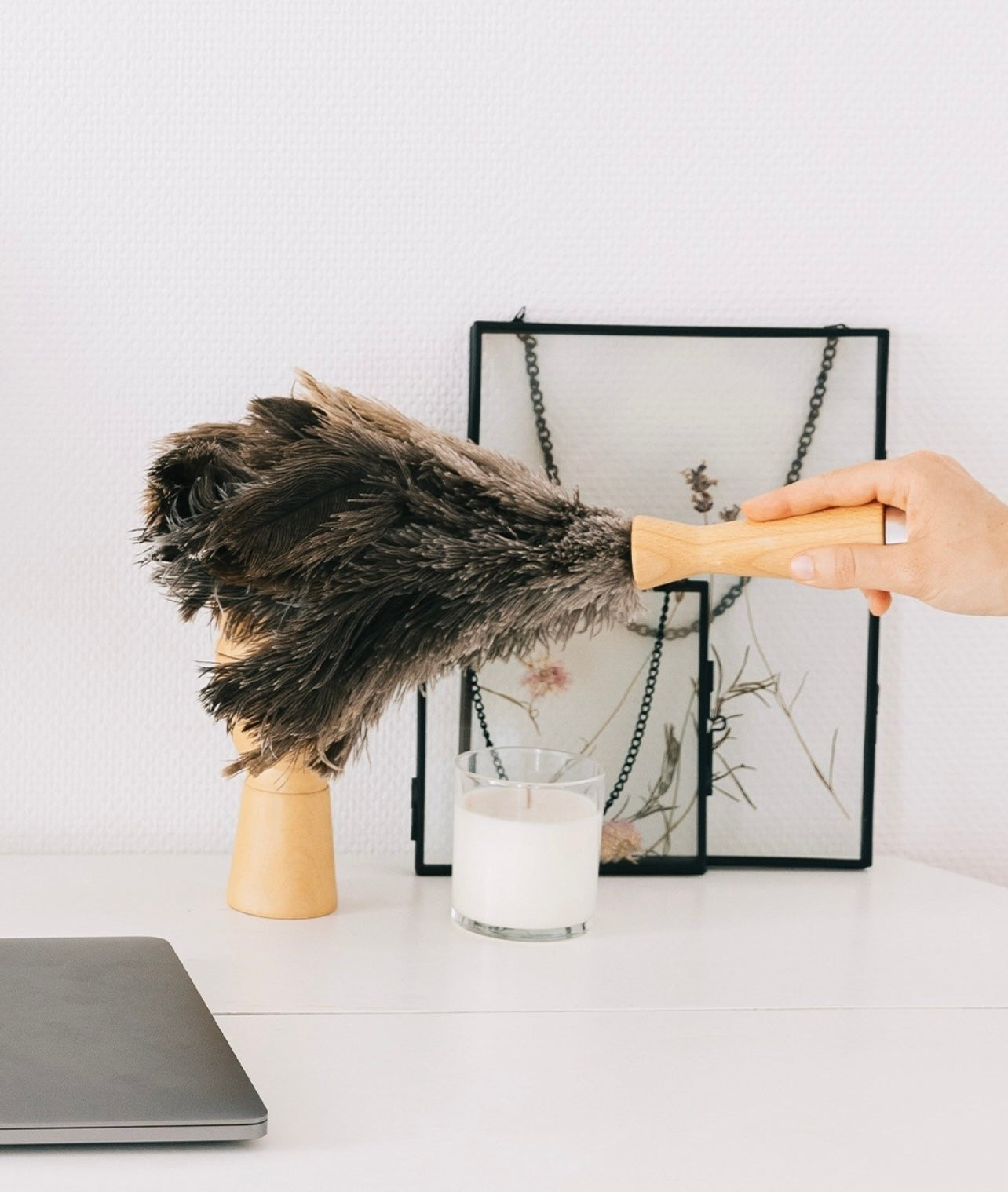 Hand holding feather duster with wooden handle next to candle, picture frame, and laptop on desk.