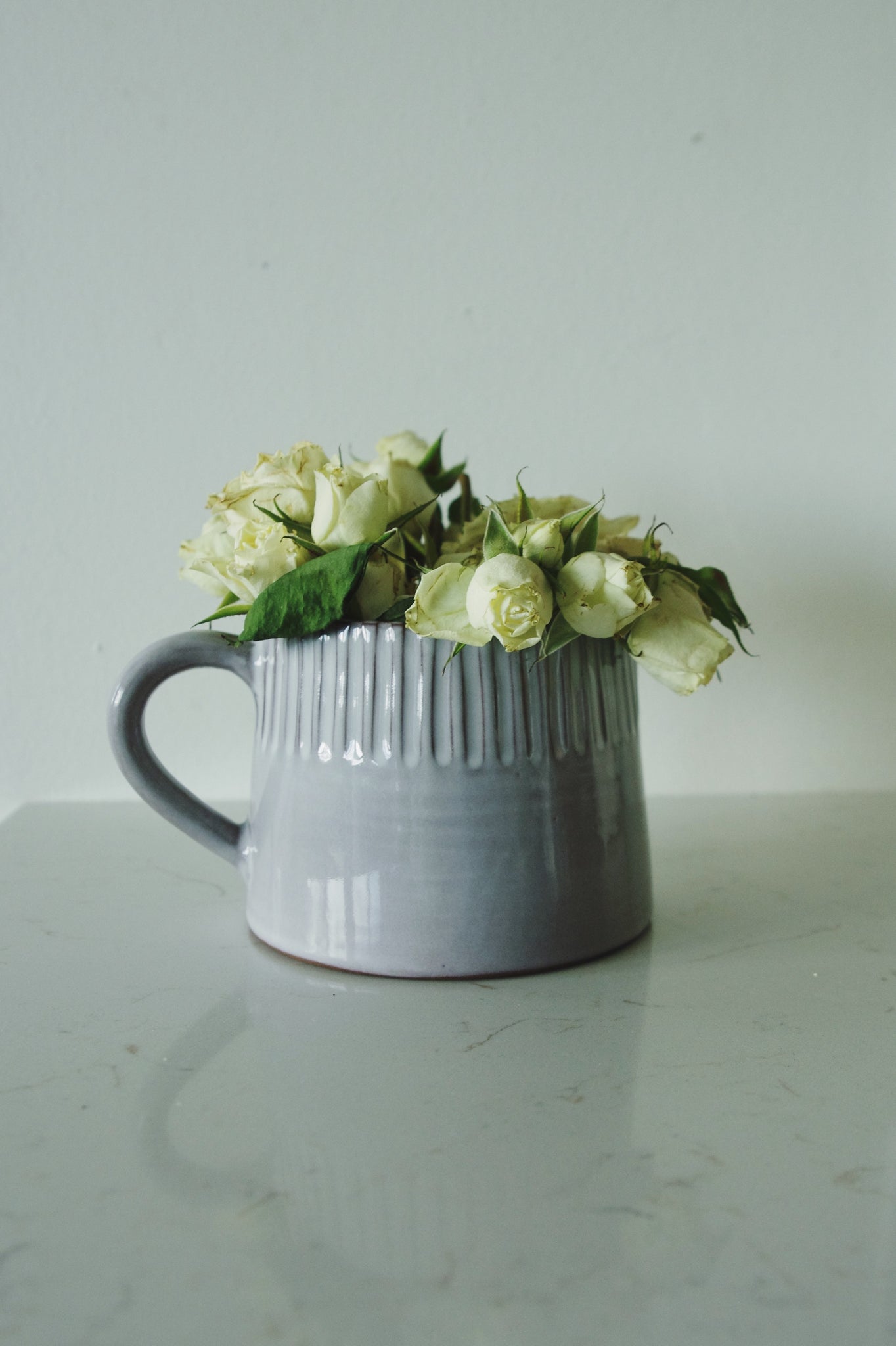 Soft grey ridged ceramic pitcher with fresh white roses on marble, against an off-white wall.