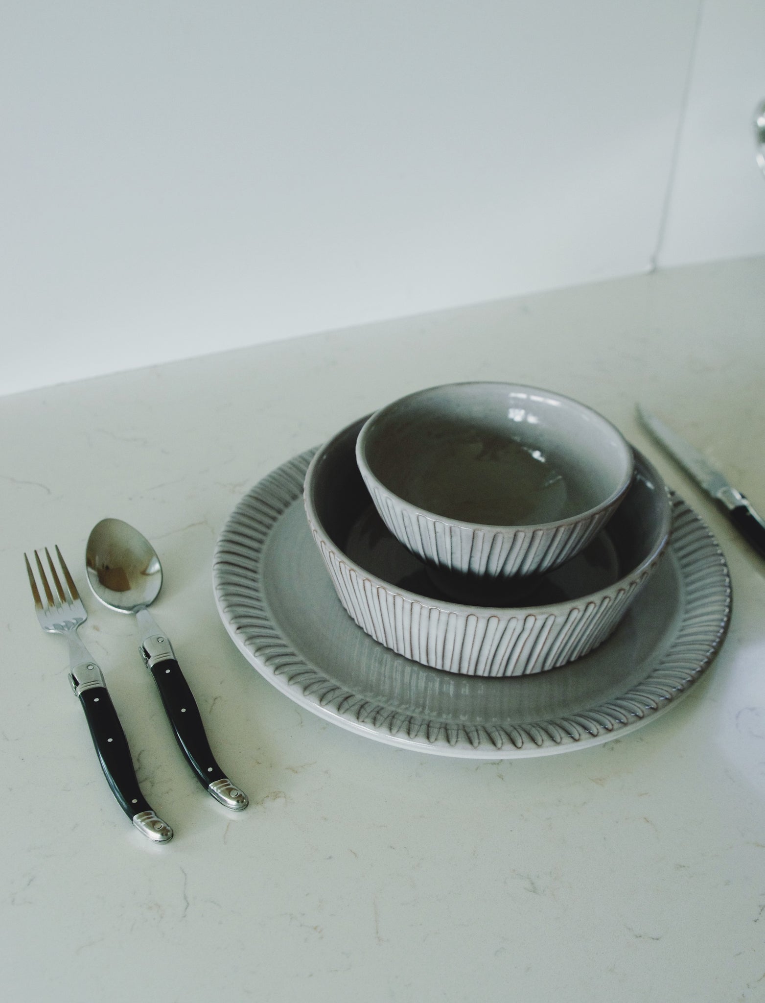 Stacked grey ribbed bowls and plate with black-handled silverware on a marble counter.