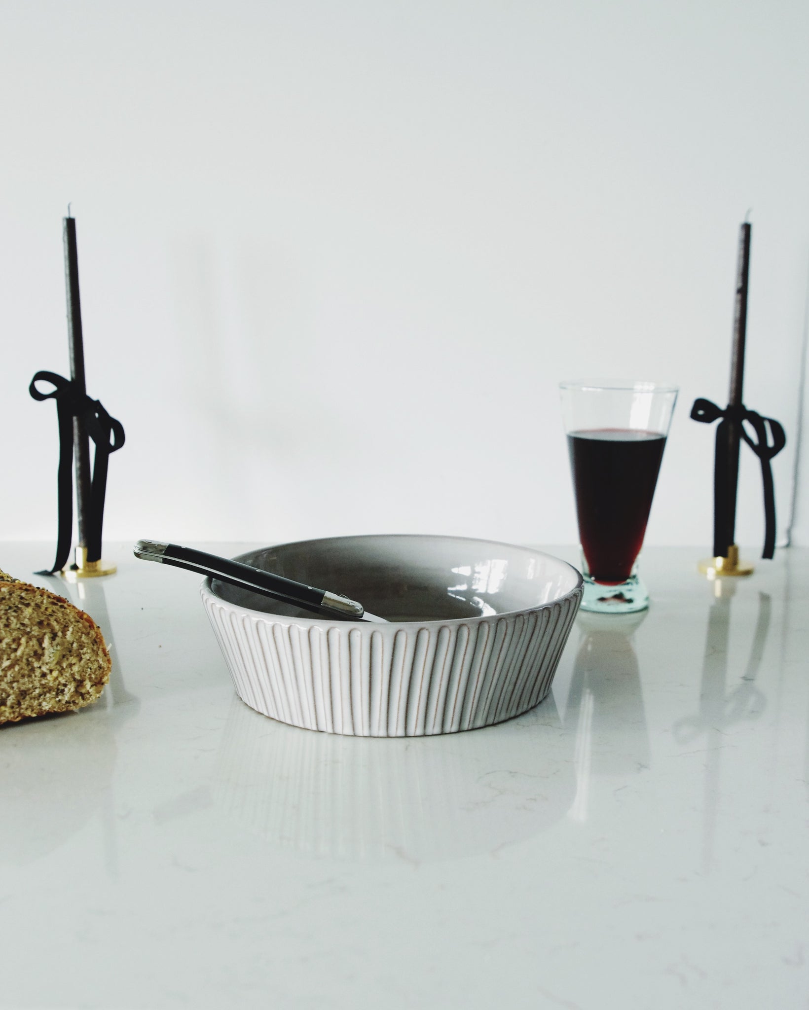 Grey ribbed pasta dish with a black-handled utensil, bread slice, and wine glass flanked by black ribbon-tied candles on marble.