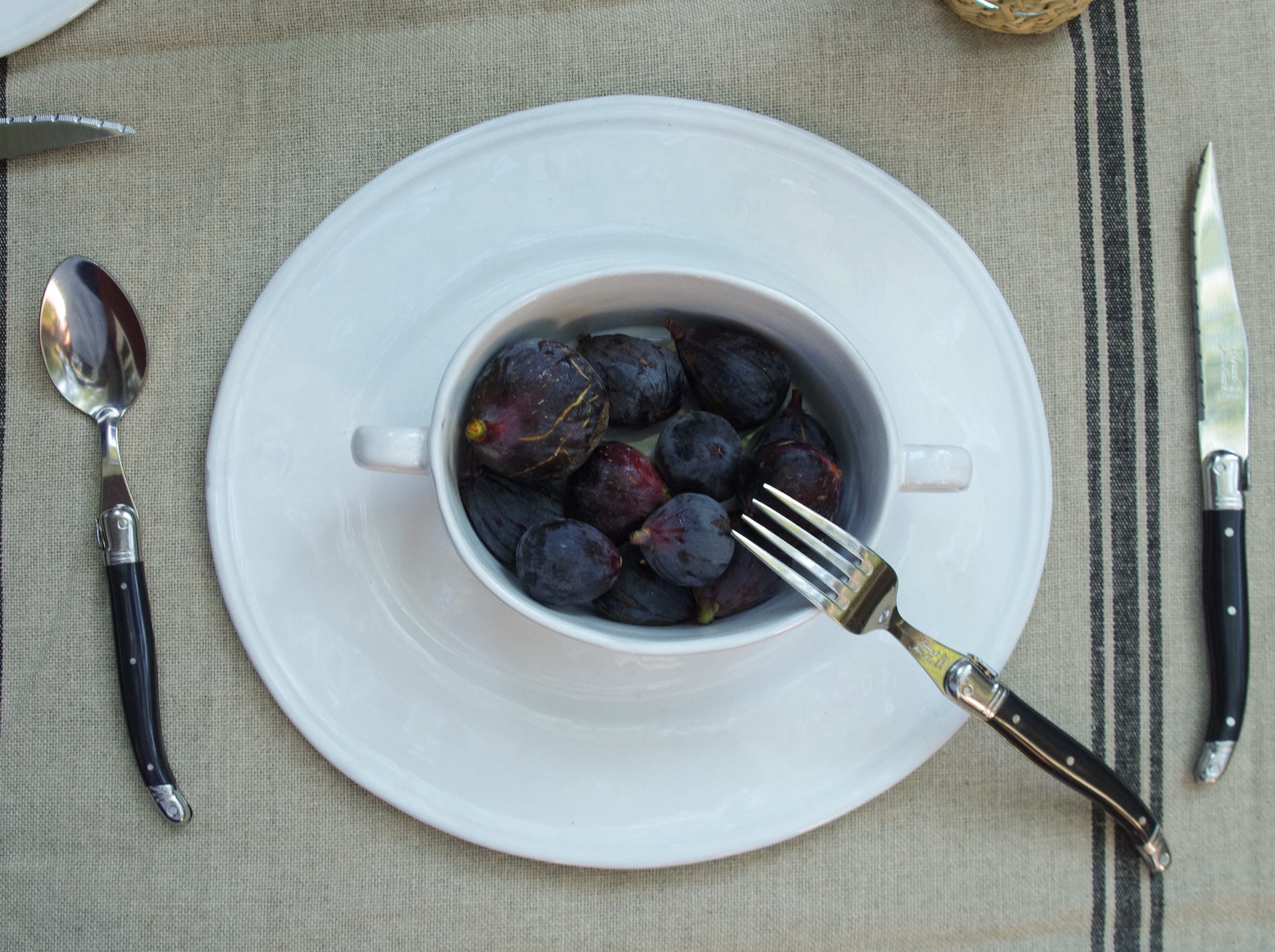 Bowl with two handles holding figs next to cutlery with black handles and large white dinner plate.