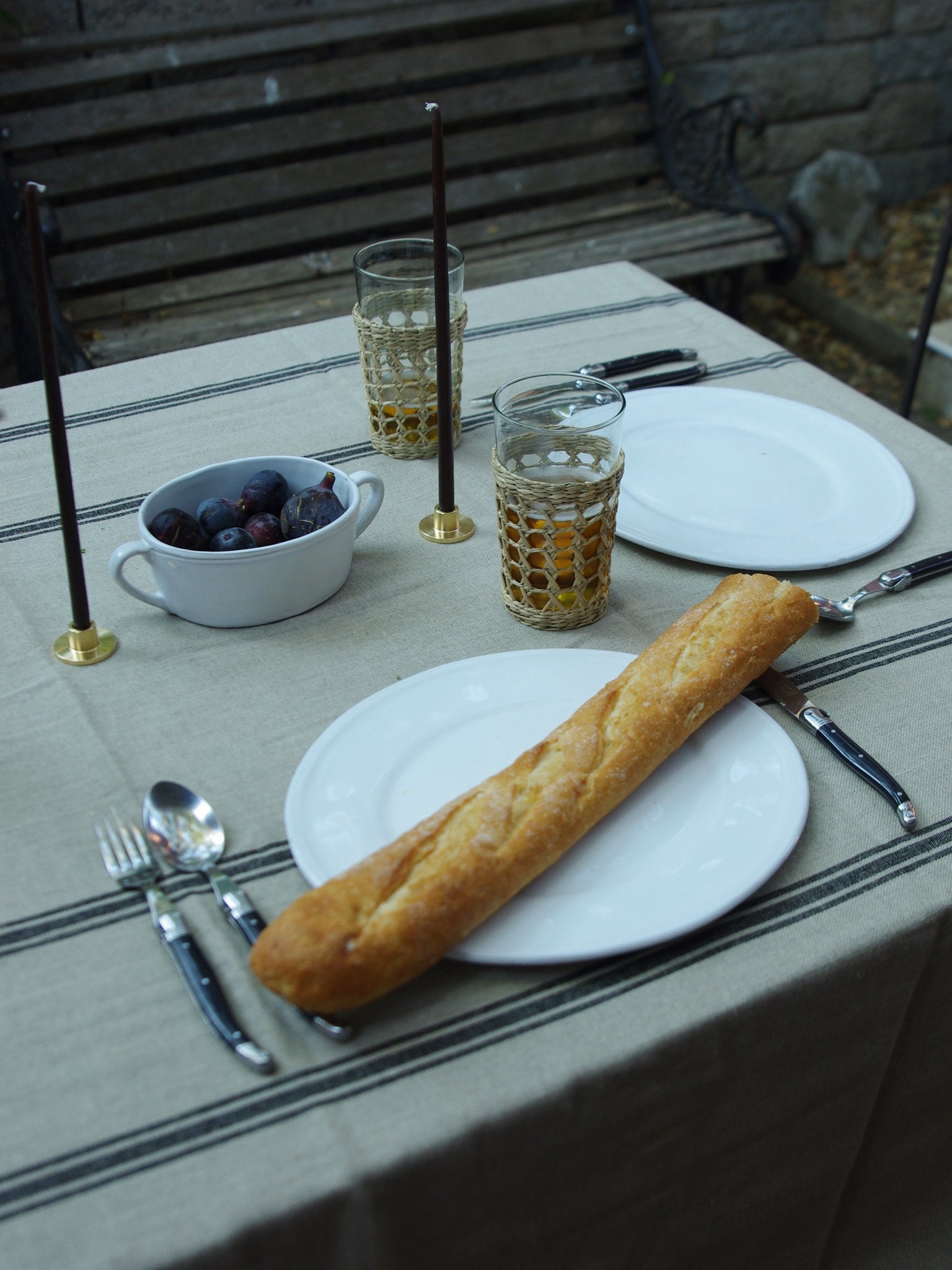 Baguette on white ceramic dinner plate. Table setting of black taper candles in gold candle holders next to black cutlery all on top of a linen tablecloth with black stripes. Outdoor setting.