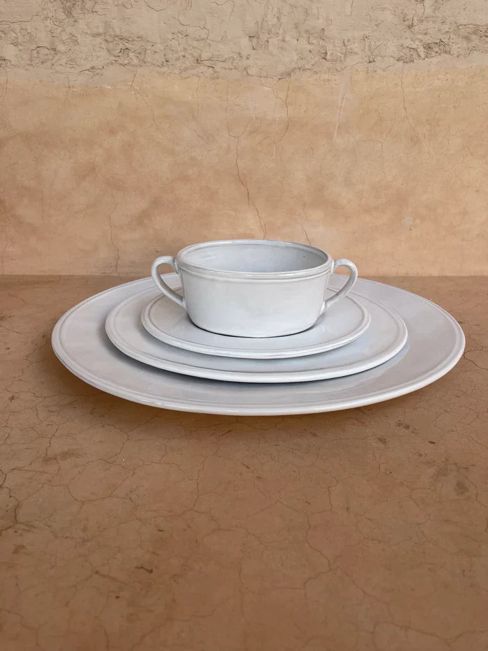Soup bowl with two handles on each side sitting on a stack of three dinner plates of various sizes.