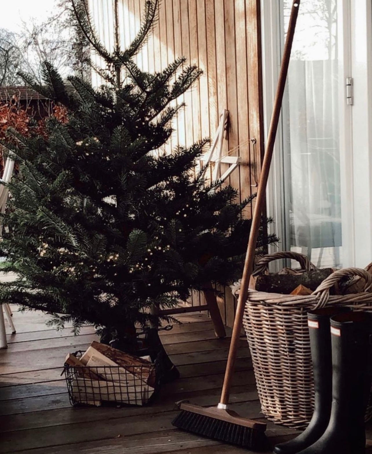 Dark Christmas tree next to leaning dark wooden broom with black bristles. Tall leather black boots sit next to a woven basket and dark wire basket holding firewood.
