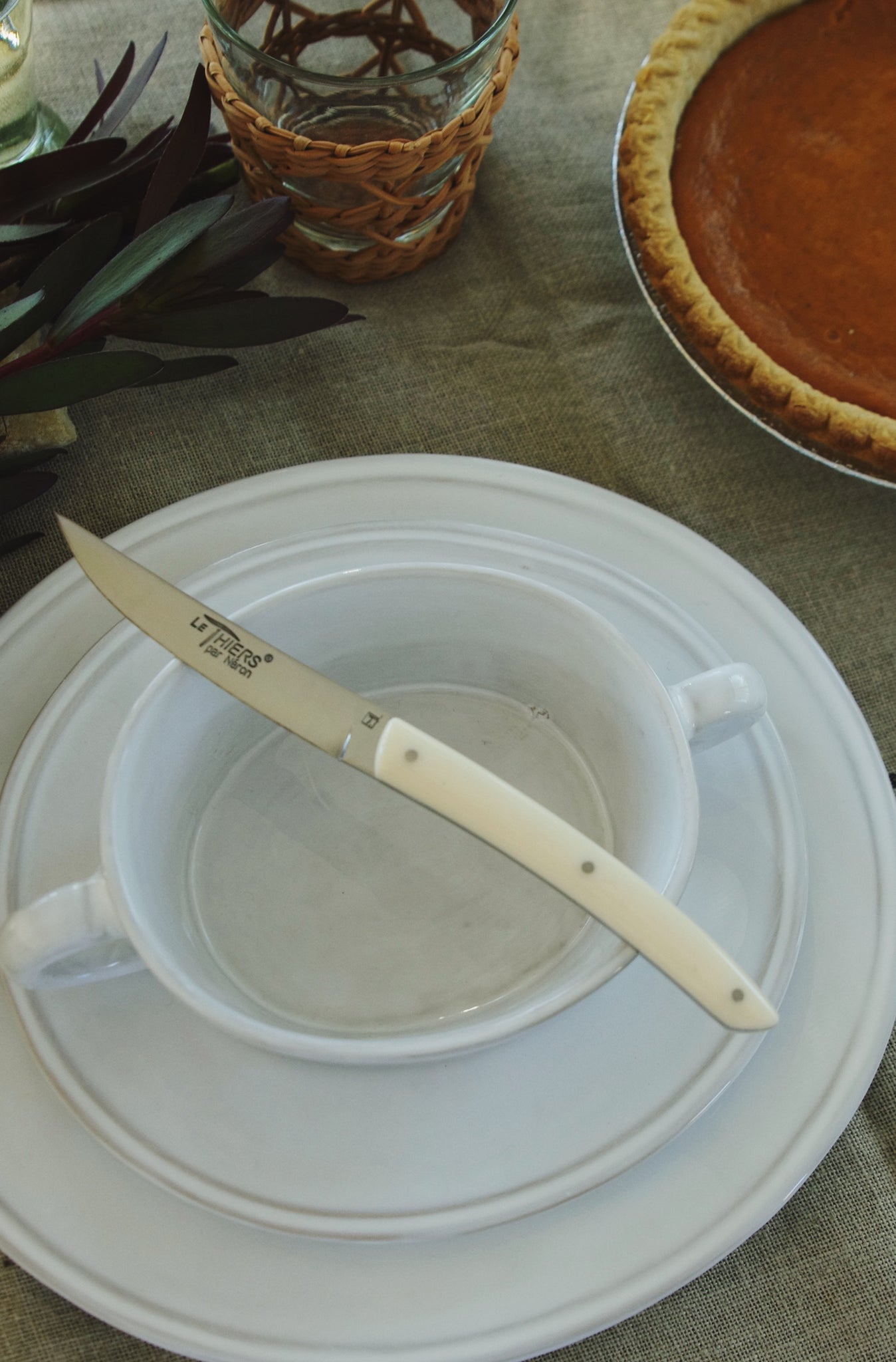 Knife with ivory color handle sitting on top of handled soup bowl which is on top of a stack of two white ceramic plates.