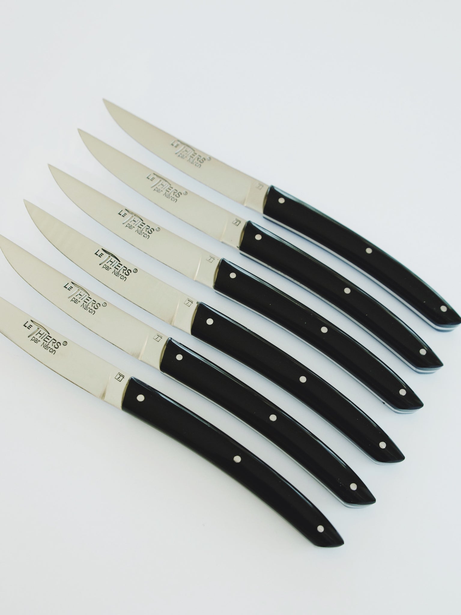 Row of six knives with black handles. Blade reads 'Le Thiers par Neron'.