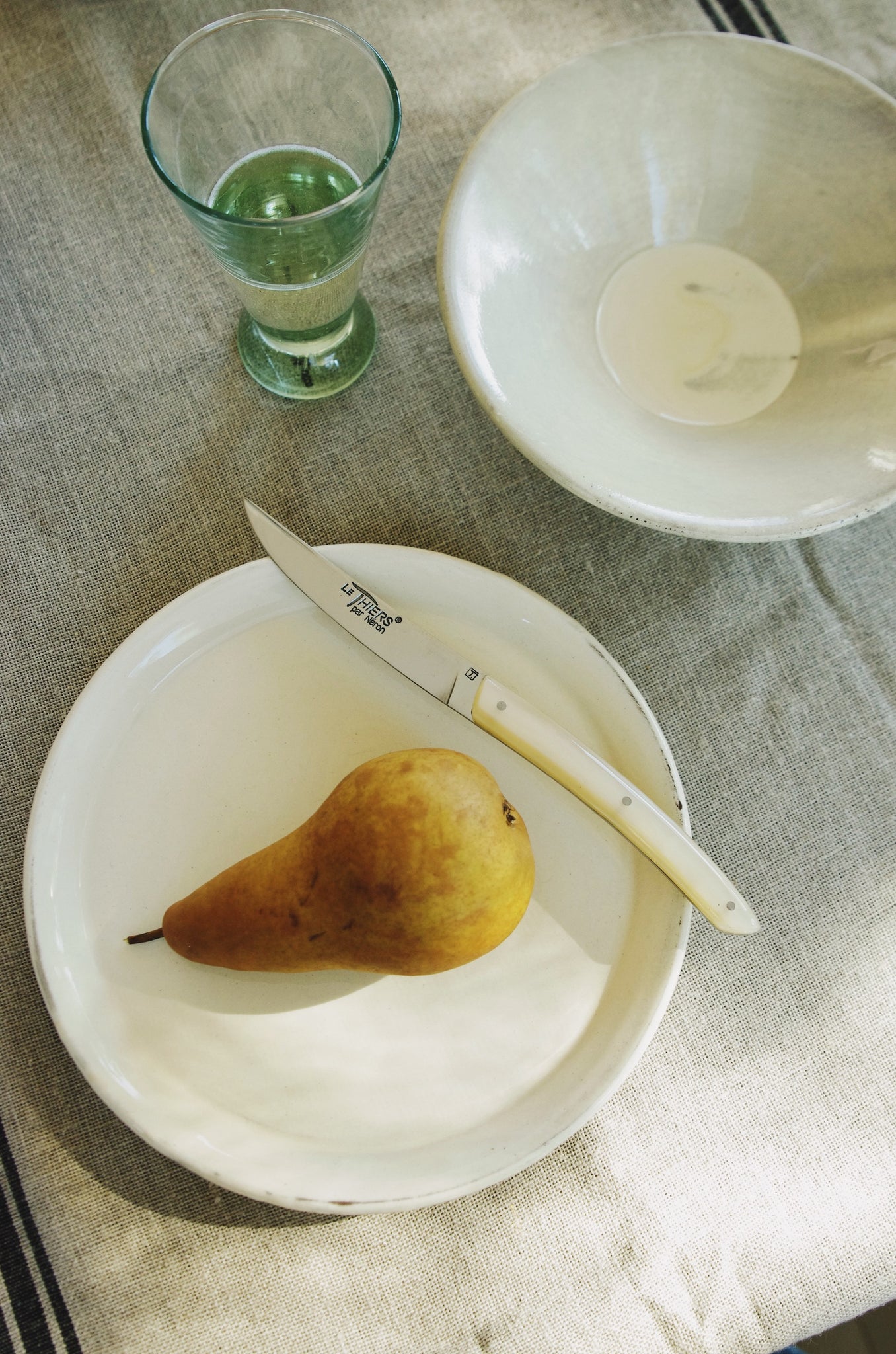 Small knife with cream handle next to pear on a rustic white plate.