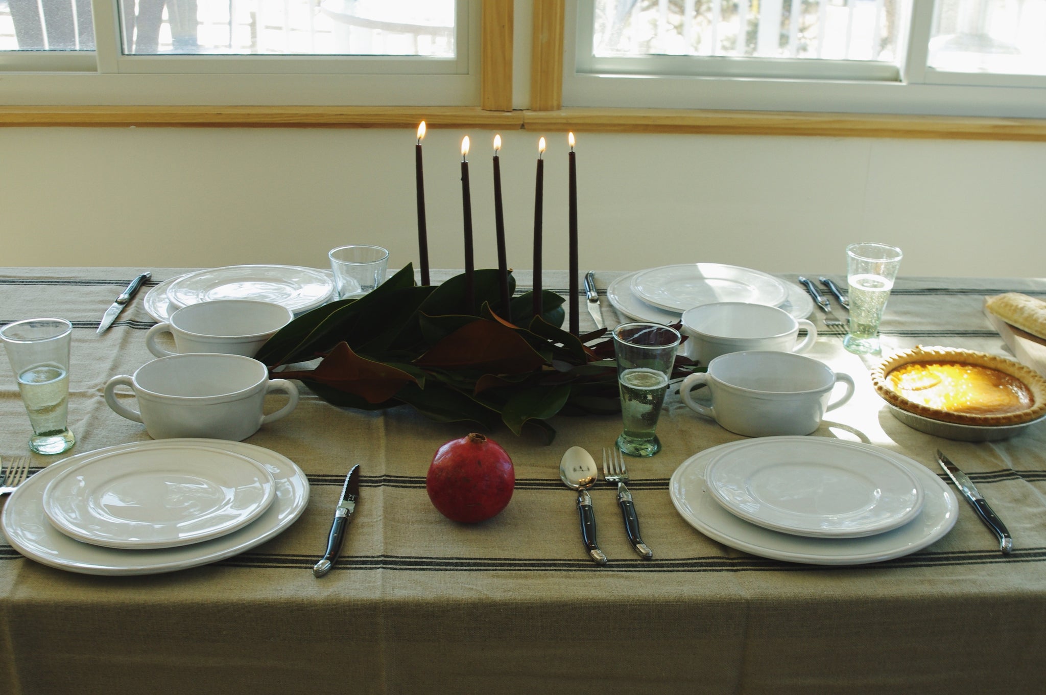 Table setting of white ceramic dinnerware, lit black taper candles, magnolia leaves garland, pomegranates, pie, small green champagne flutes, and black cutlery.