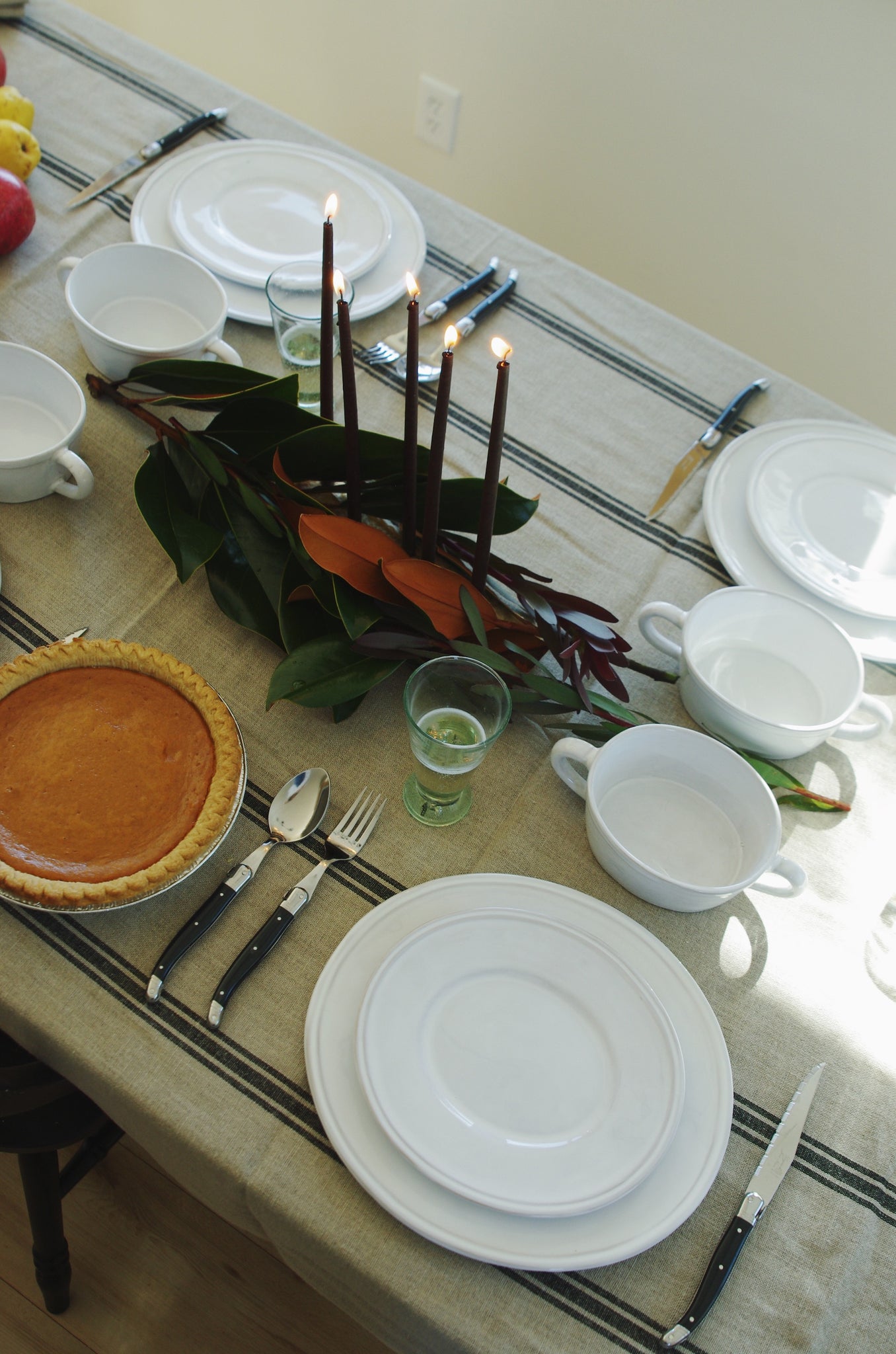 Linen tablecloth with black stripes. On the table are white ceramic plates and bowls. Magnolia leaf garland next to pie.