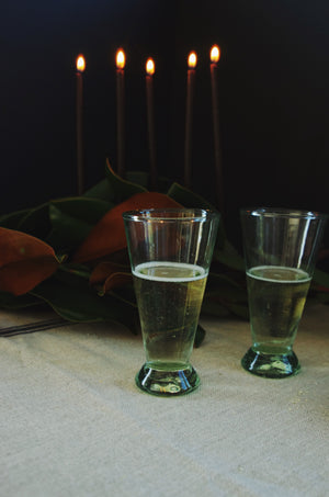 Two small green glasses next to magnolia leaves and black candles on linen tablecloth.