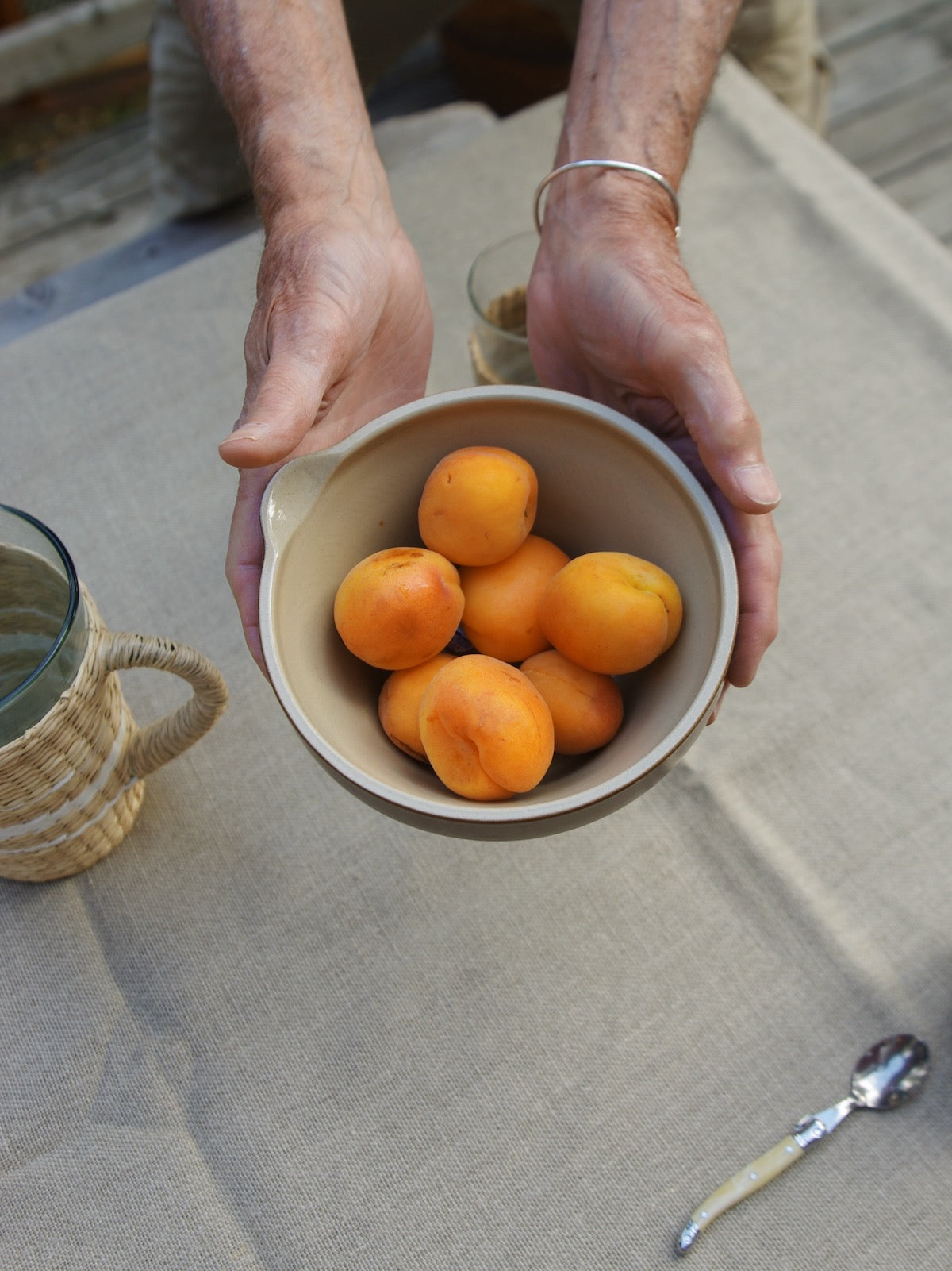 Hands holding bowl with pour spout full of orang fruit. Table covered with linen table cloth with seagrass pitcher and cream coffee spoon on table.