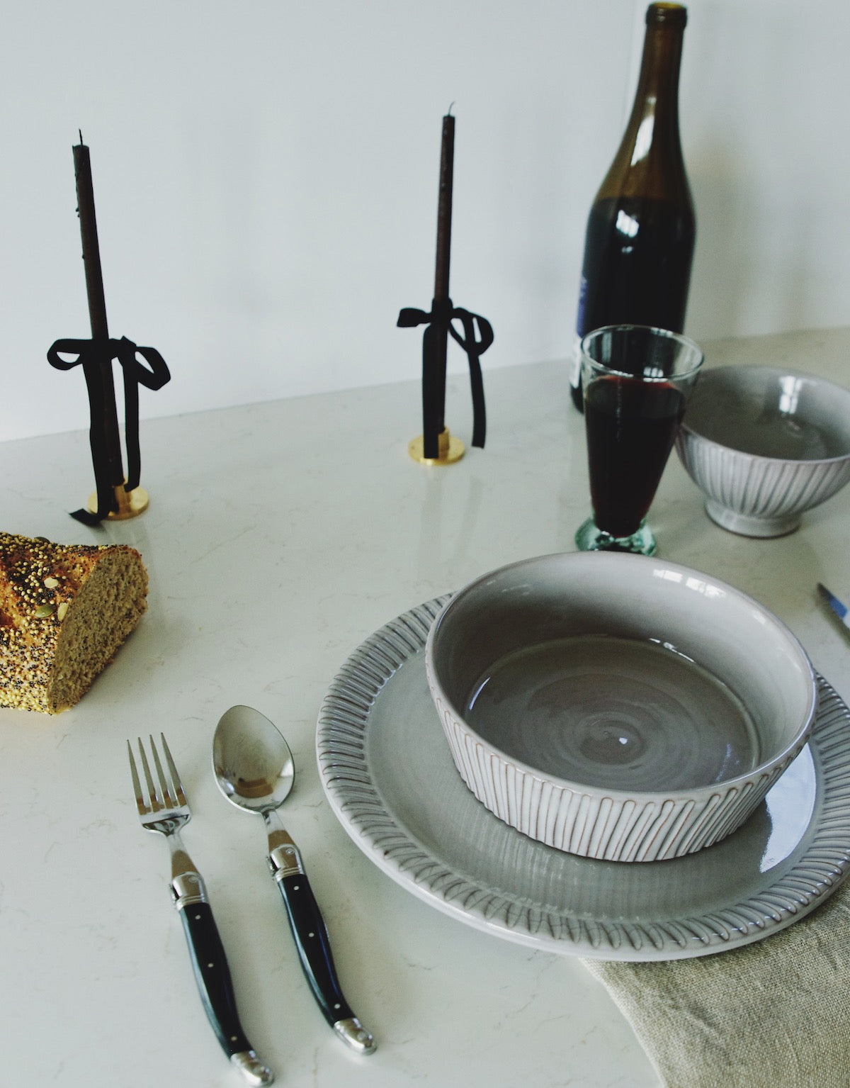 A stylish table setup with French charm, showcasing a gray ribbed dinner plate topped with a matching bowl. Beside the plate lies a set of French Laguiole cutlery with black handles, including a spoon and fork. In the background, two black candles with ribbon-tied brass holders, a half-eaten loaf of seed-encrusted bread, a bottle of red wine, and a filled wine glass add to the ambiance, all set on a marble countertop.