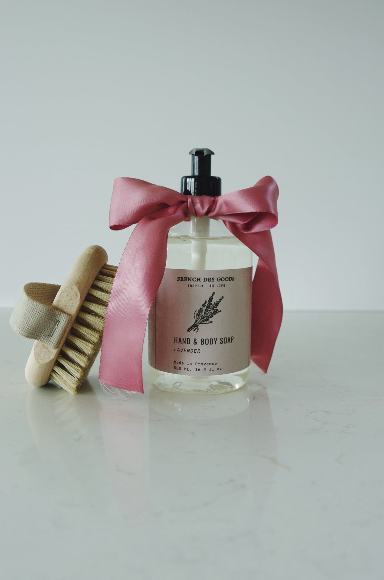 Liquid hand and body soap pump bottle labeled "Lavender", tied with a pink satin ribbon, next to a wooden nail brush.