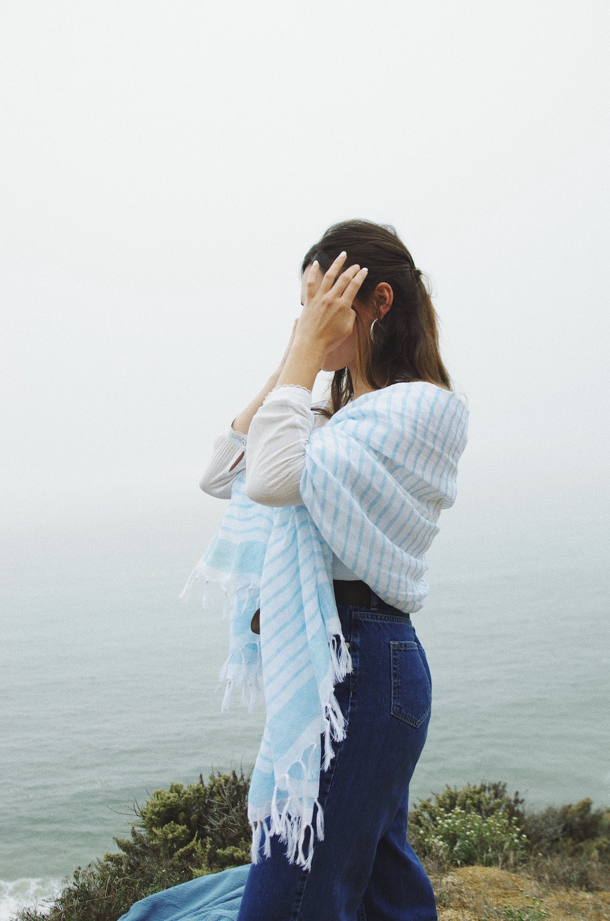 A woman with her back turned to the camera, wearing a white blouse and blue jeans, has a blue and white striped scarf over her shoulder. Her hand is touching her hair, and she's overlooking a foggy coastal landscape.