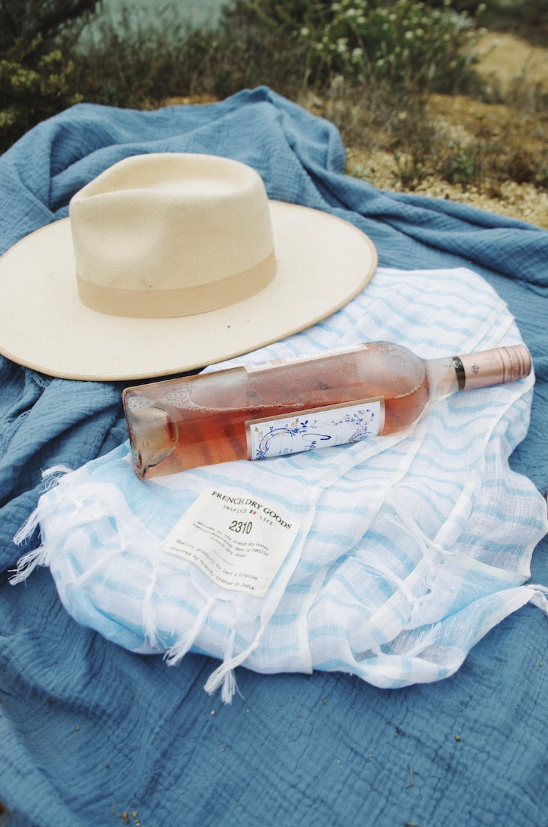 A beige wide-brimmed hat and a bottle of rosé wine lie on top of a striped blue and white scarf, which is spread out over a textured blue blanket amidst a natural, grassy background.