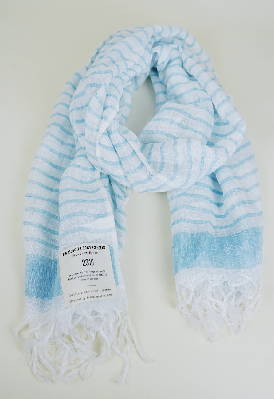 A light blue and white striped scarf with frayed tassels on the ends, showcasing a label that reads 'FRENCH DRY GOODS 2310 INSPIRED LIFE,' is neatly laid out on a white background.