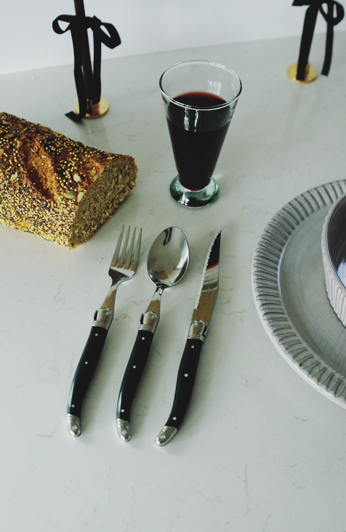 An elegant table setting featuring a selection of French Laguiole cutlery with black handles, including two knives, a fork, and a spoon, arranged to the left of a gray ceramic plate with ribbed edges. A slice of multi-seed bread, a glass of red wine, and a candle with a black bow tied around its holder complete the sophisticated dining atmosphere, all resting on a marble surface."