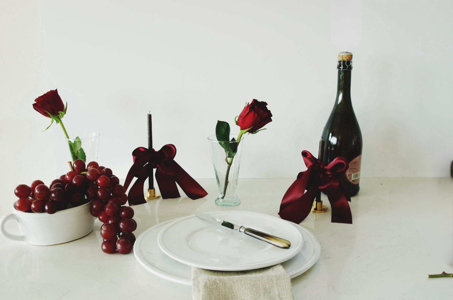 Elegant table setting featuring a cluster of red grapes in a white bowl, a single red rose in a clear glass, and a wine bottle with a dark red ribbon, alongside stacked white plates with silver cutlery on a linen napkin, all on a white surface against a white wall.