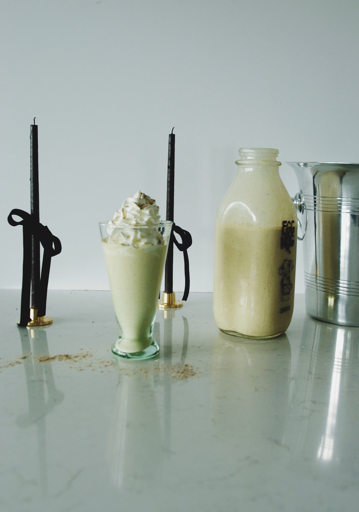 Milkshake with whipped cream in a glass flute, vintage milk bottle, metal pitcher, and ribbon-tied candles on marble.