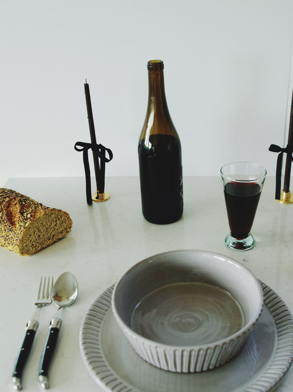 Table setting with grey ceramic dishes, black-handled silverware, a slice of bread, wine glass, bottle, and candle holders on a marble surface.