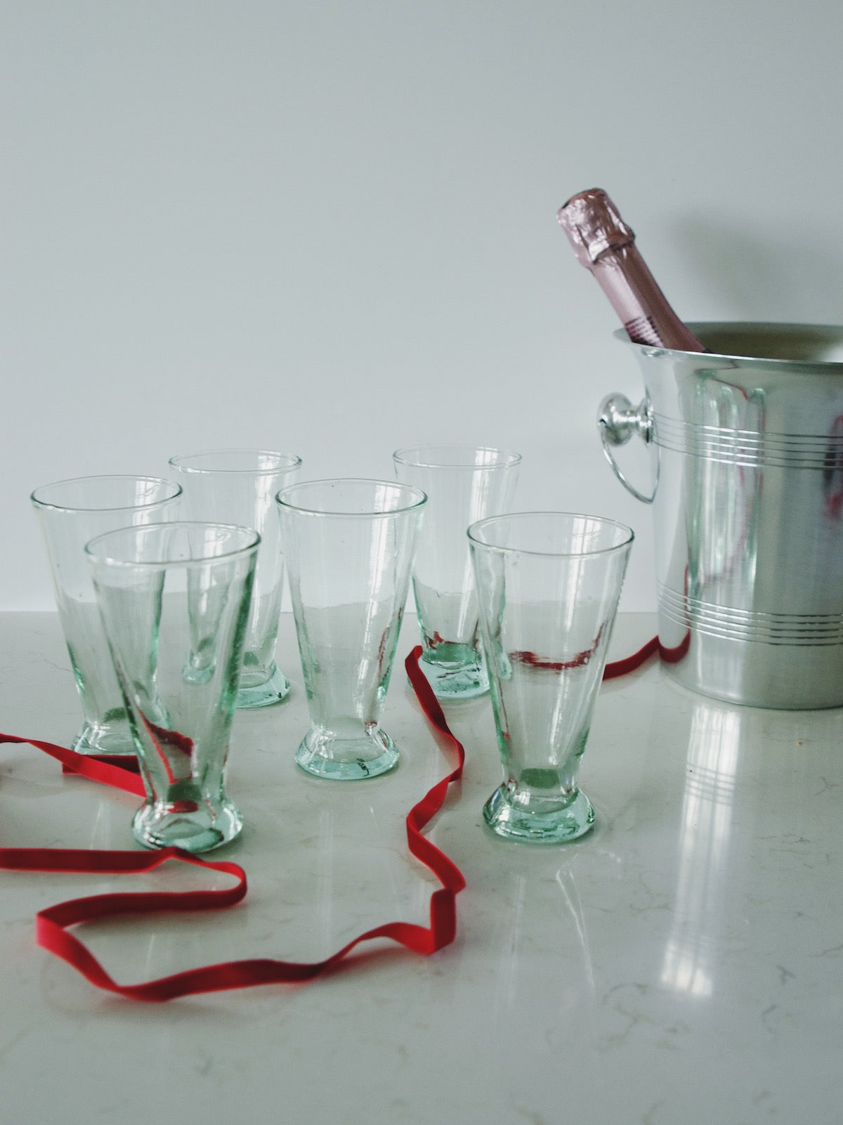 Recycled wine flutes with a red ribbon and champagne bottle in a metal ice bucket on a marble surface.