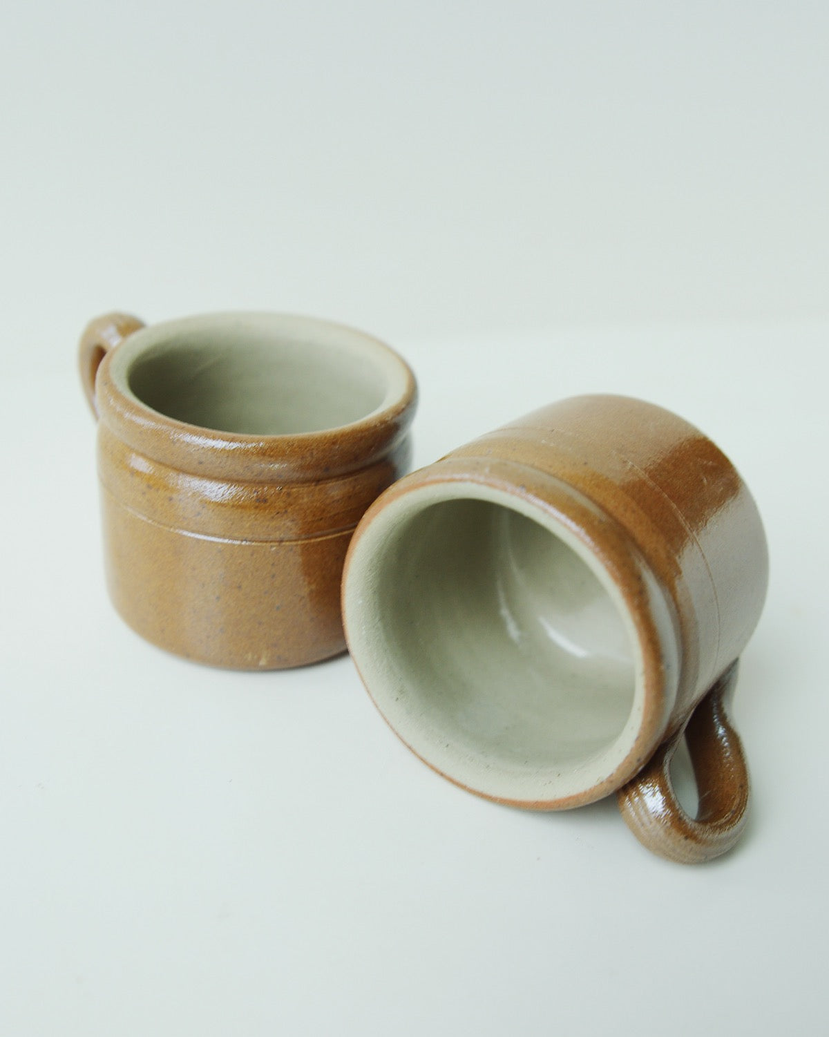 Two ceramic mugs with handles, one standing upright and the other tipped on its side, showcasing their glazed interior. 