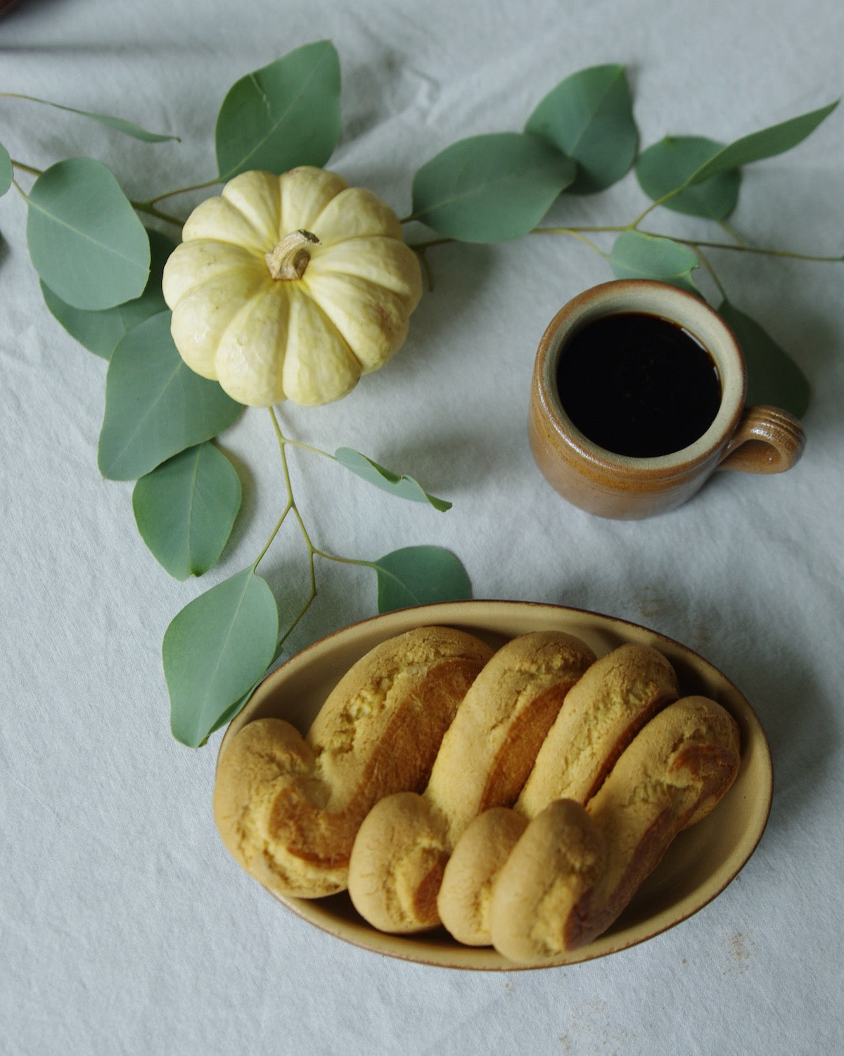 Coffee mug next to pumpkin, eucalyptus leaves, and bowl of pastries.