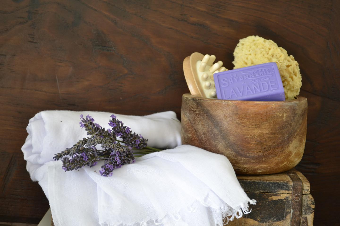 Purple bar of soap reading La Savonnerie de Nyons Lavande in wooden bowl along with bath body scrubbers and sponge, next to white towel with lavender branches strewn on top.