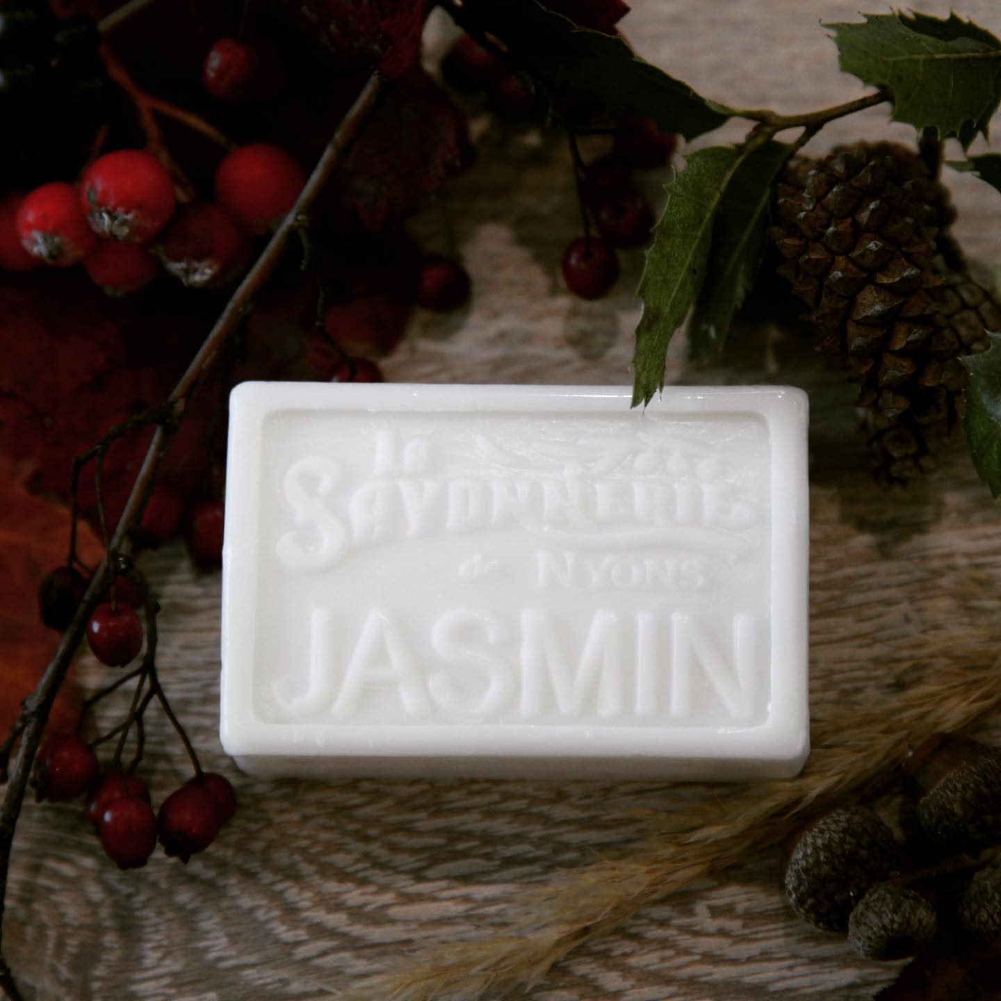 White bar of soap with stamp reading La Savonnerie de Syons Jasmin surrounded by holly berries and pinecones.
