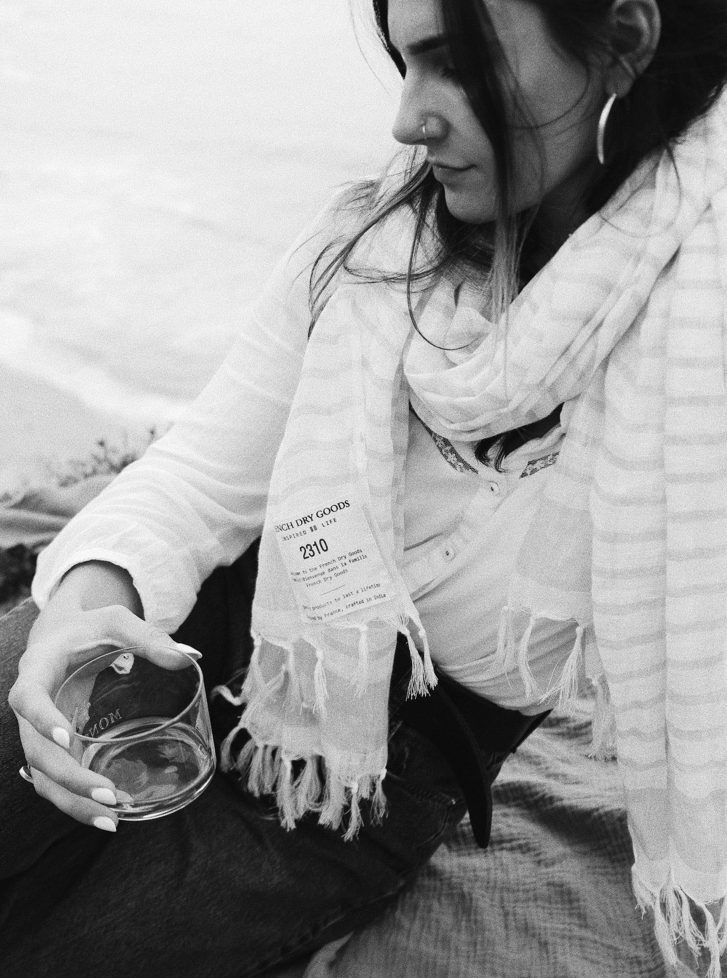 A black and white image of a seated woman in a white blouse and jeans, holding a clear glass in her hand. She is wearing a striped scarf with a visible label and tassel edges, looking downwards.