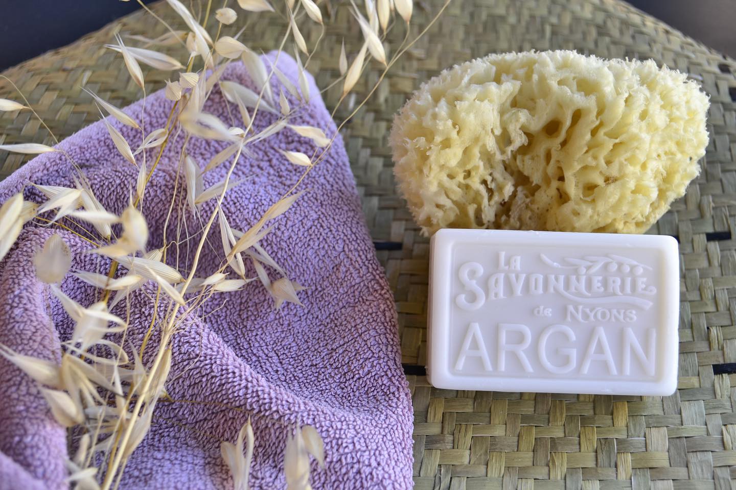 White bar of soap that reads La Savonnerie de Nyons Argan next to a natural bath sponge, sprigs of wheat, and a purple towel.