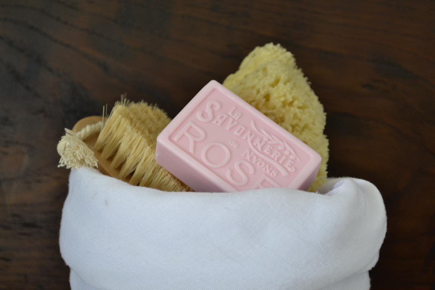 Pink bar of soap with the words La Savonnerie de Nyons Rose on it, wrapped in a towel along with a body scrub brush and sponge.