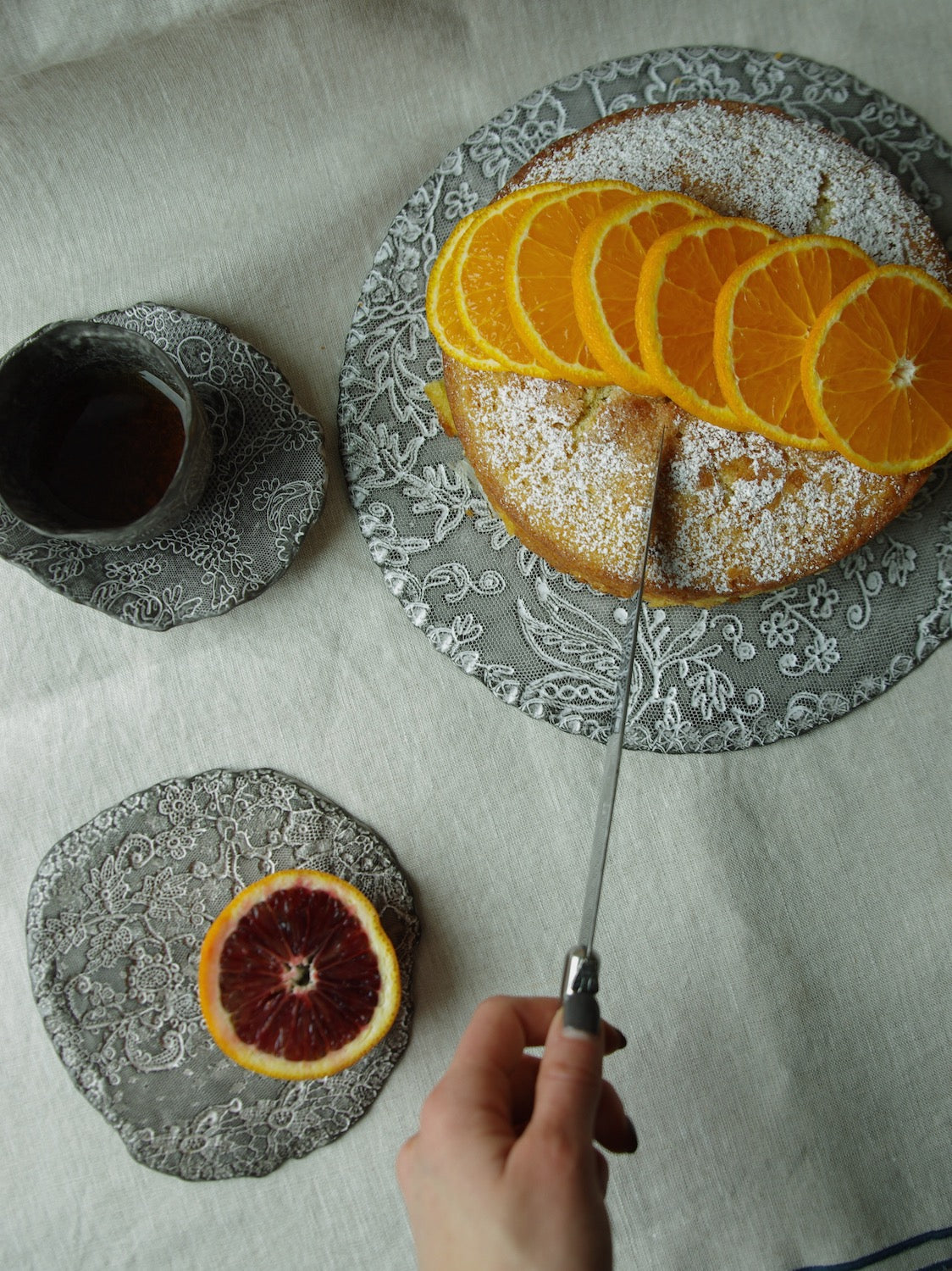 Cake covered in orange slices served on lace pattern ceramic platter with Laguiole cake slicer.