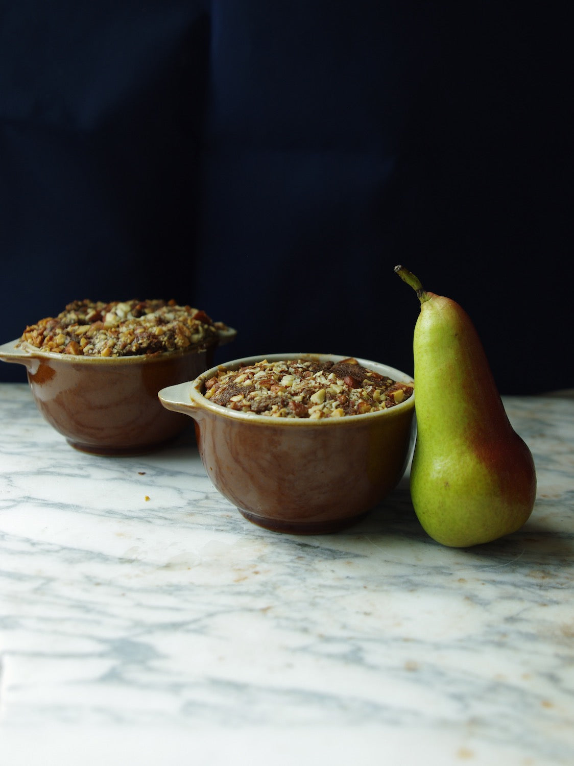 Two brown handled bowls holding baked nut bread next to a pear on a marble counter.