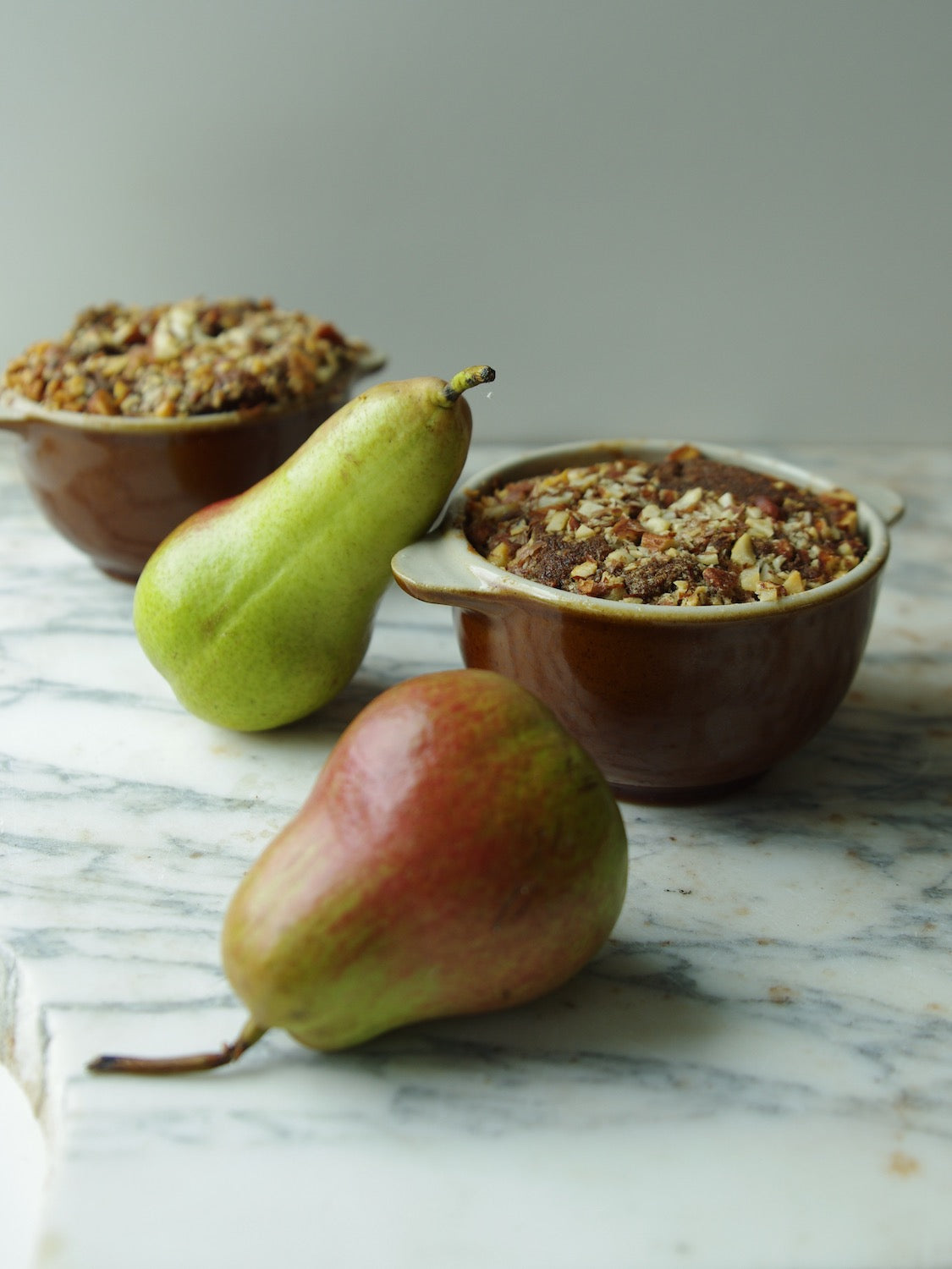Two brown handled bowls holding baked nut bread next to two pears on a marble counter.